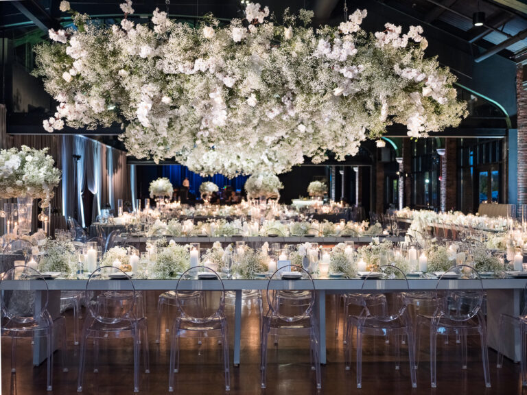 All-white wedding reception with massive suspended floral cloud installation, ghost chairs, and long banquet tables with candles