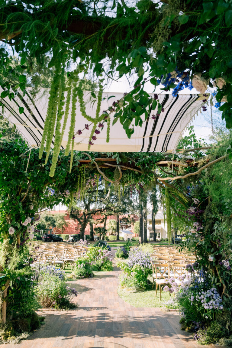 Outdoor garden wedding ceremony entrance with lush greenery arch, purple and blue floral arrangements, striped canopy, and cross-back chairs