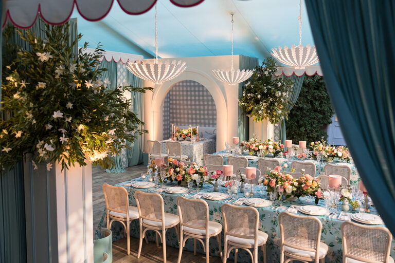 Elegant tented wedding reception with blue floral linen, pink rose centerpieces, shell chandeliers, and arched gingham backdrop