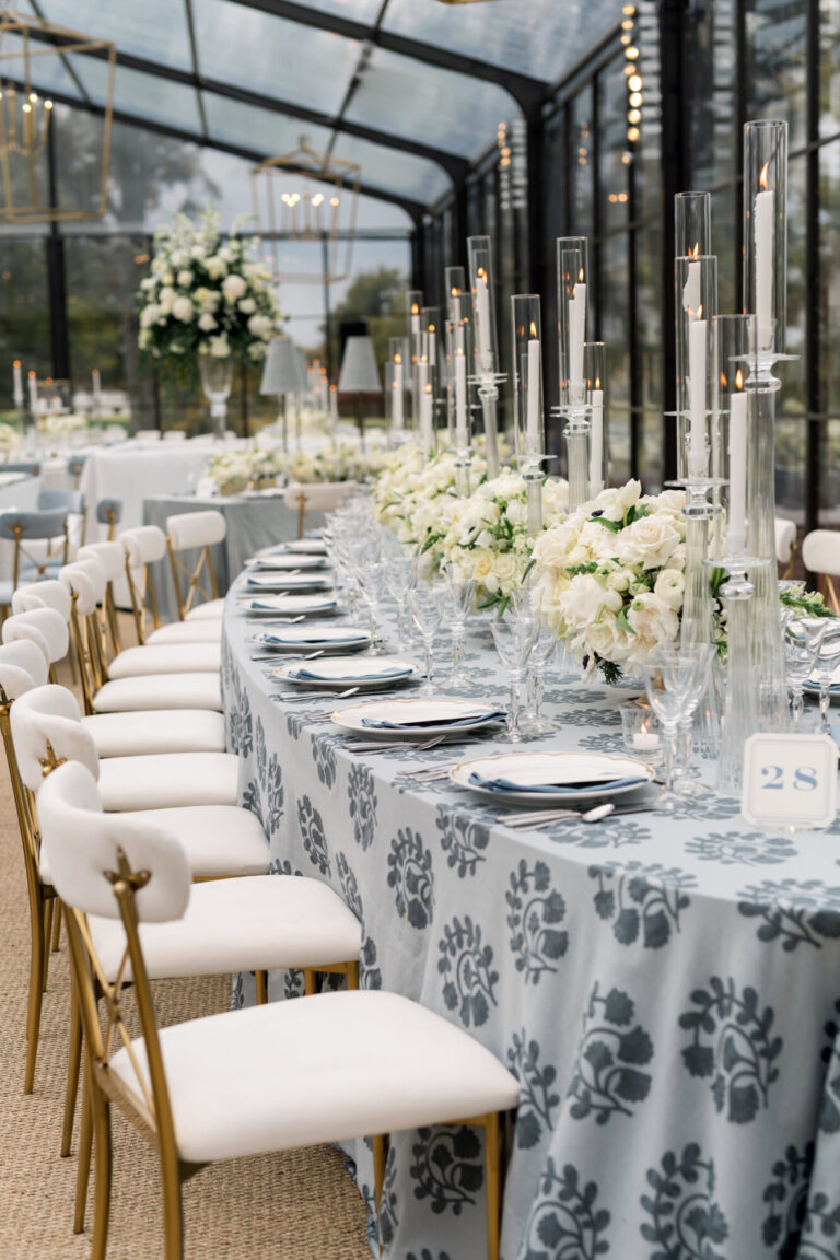 Luxury wedding reception long table with blue floral patterned linen, white rose centerpieces, and tall crystal candlesticks in a glass greenhouse venue