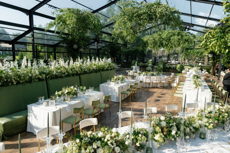 Luxury greenhouse wedding reception with white and green floral installations, green velvet banquette seating, and tall taper candles