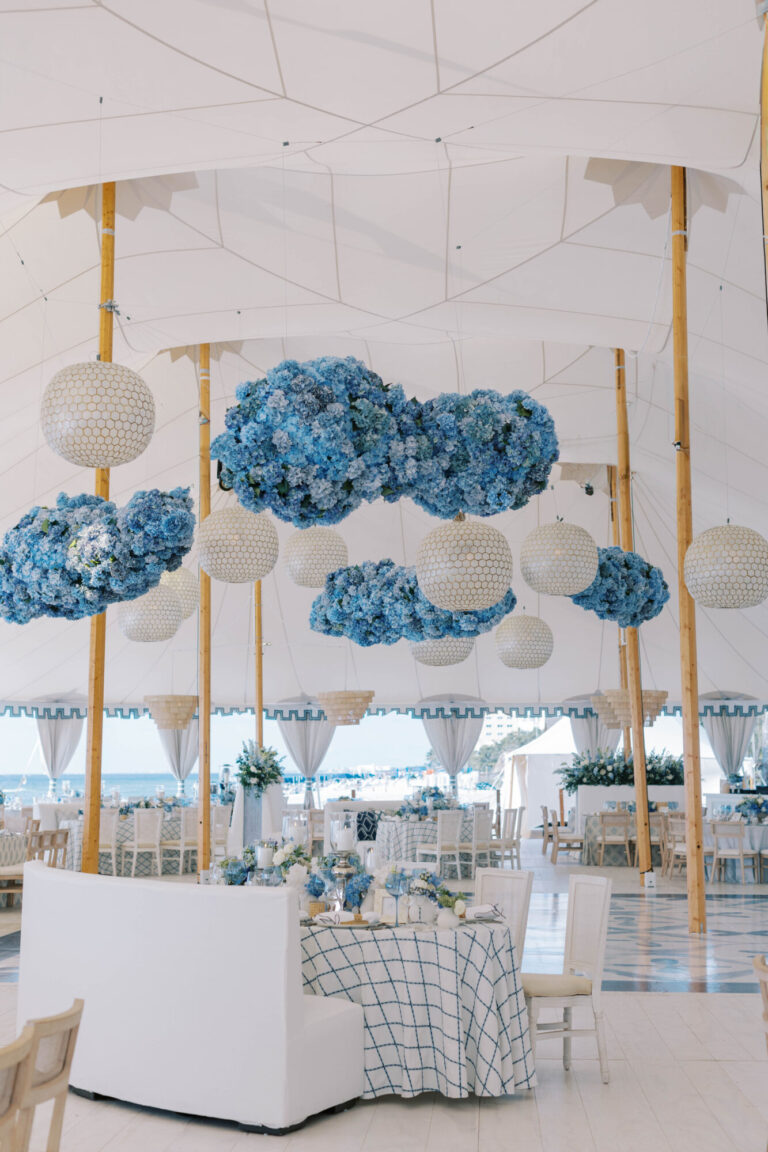 Destination beach wedding reception tent with suspended blue hydrangea cloud installations and white globe pendant lights over dining tables