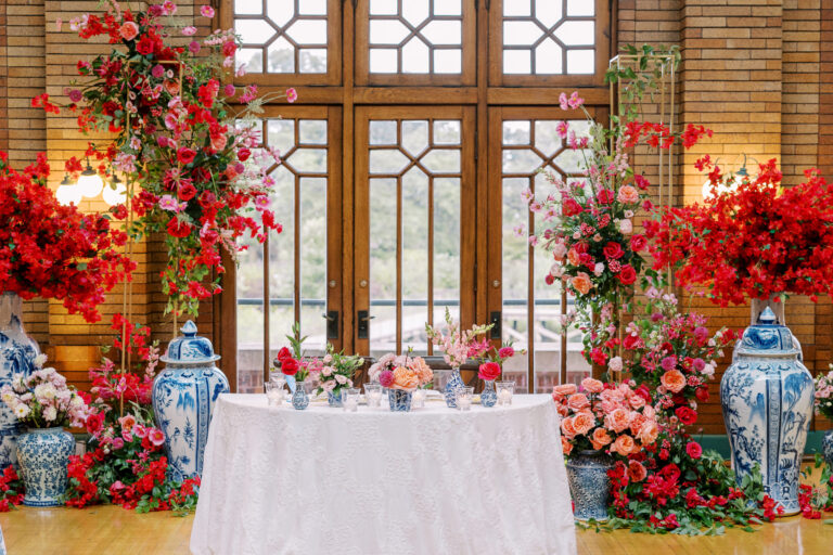 Wedding sweetheart table surrounded by dramatic red and pink floral installations and large blue and white ginger jar vases in a brick venue