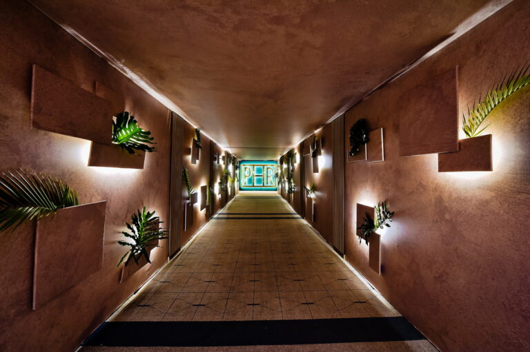 Dramatic Bar Mitzvah entrance hallway with terracotta textured walls, tropical plant wall installations, warm uplighting, and glowing neon monogram sign at the end