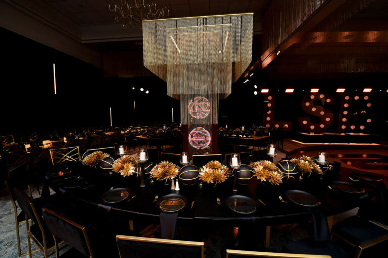 Dramatic basketball themed Bar Mitzvah reception with black and gold table settings, gold dahlia centerpieces, crystal chain chandelier, and illuminated marquee letters on stage