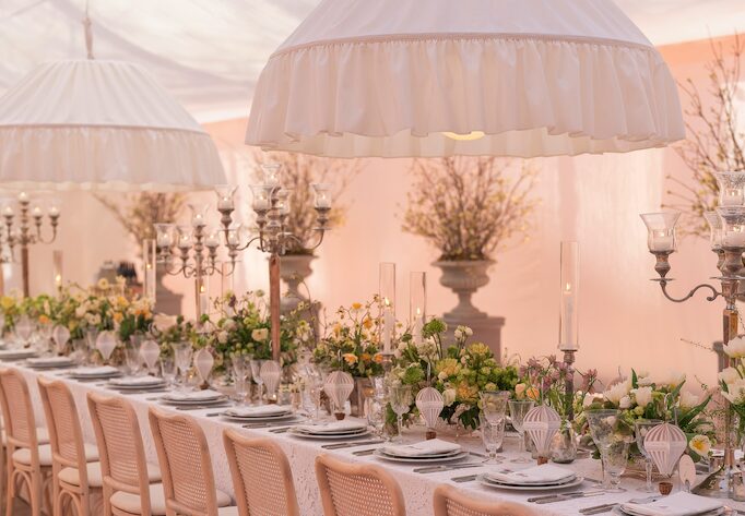 Long banquet table under ruffled white canopy lights, set with blush cane chairs, silver candelabras, and soft pastel floral centerpieces